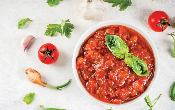 A small bowl filled with home-made passata, topped with basil leaves, and surrounded by garlic, tomatoes, and parsely.