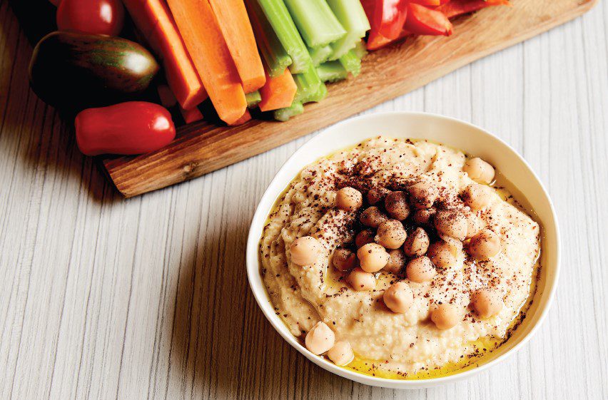 A small bowl of hummus with brightly coloured vegetables, sitting on a chopping board.
