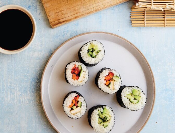 A plate with colourfully filled nori rolls, with pot of rice and bamboo rolling mat.