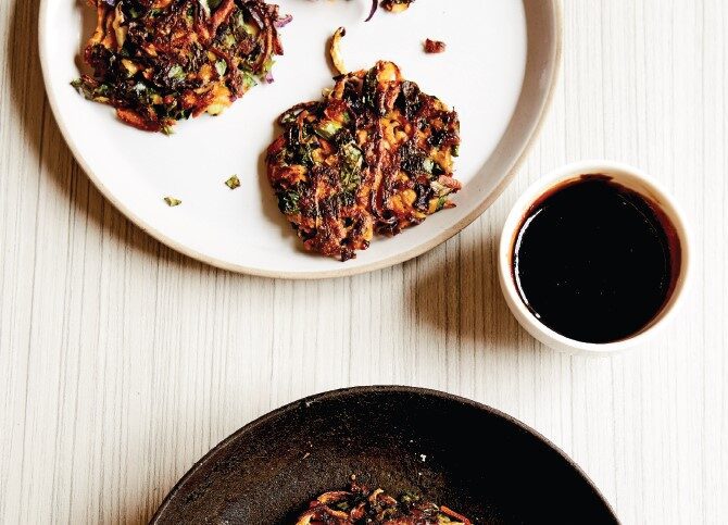 A plate and frypan with three crispy-looking grated vegetable pancakes, with soy dipping sauce on the side.