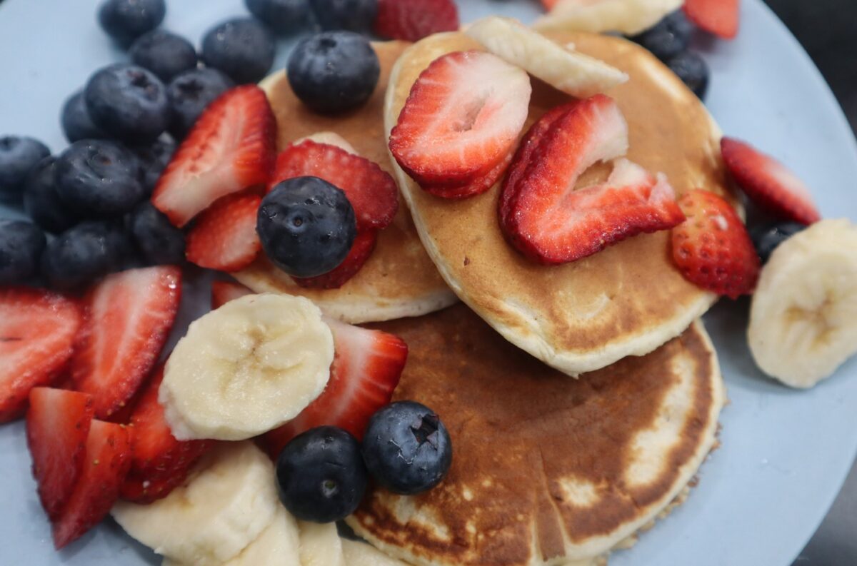 A plate with three pancakes, drizzled with honey, topped with blueberries, strawberries, and banana.
