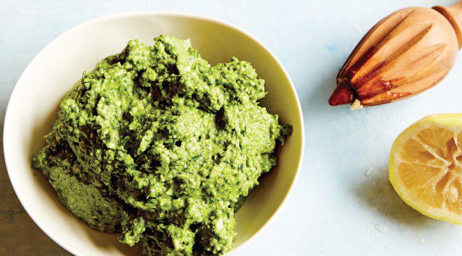 Vivid green pesto in a small bowl, next to a freshly squeezed lemon and wooden lemon juicer tool.