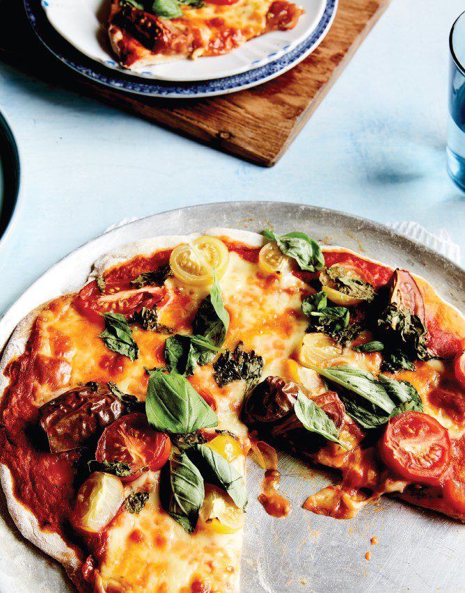A pizza, fresh out of the oven, layered with cherry tomatoes and basil, with cheese oozing onto tray.