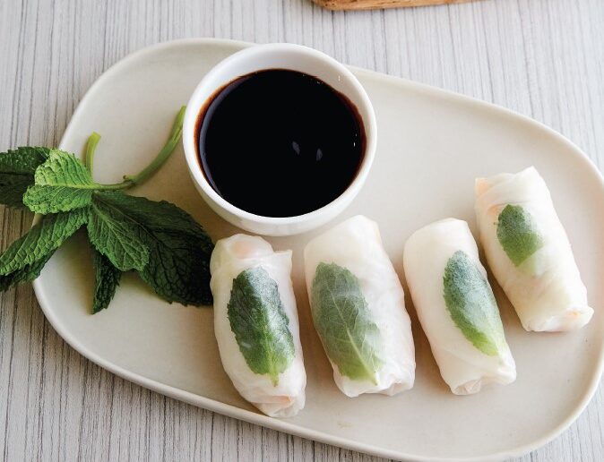 A platter of four rice paper rolls and soy dipping sauce, next to a wooden chopping board with strips of vegetables.
